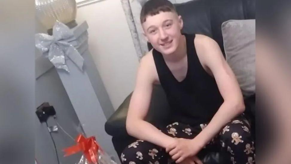 A teenage boy smiles at the camera at Christmas. He is wearing pyjama bottoms with gingerbread men on, and a black vest. He has short hair. 