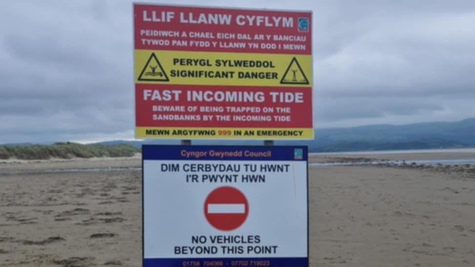 Arwyddion ar y traeth yn dweud 'Dim cerbydau tu hwnt i'r pwynt hwn' a 'Llif llanw cyflym, peidiwch a chael eich dal ar y banciau tywod pan fydd y llanw yn dod i mewn'.