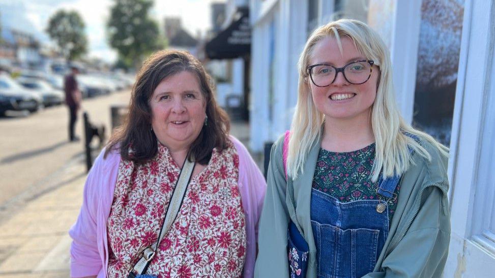 Kay and Sonnie Mead are looking at the camera standing in a street. Kay has shoulder length dark hair and is wearing a patterned pink top and cardigan. Sonnie has long blond hair and is wearing dark-rimmed glassed, dungarees and a blue jacket
