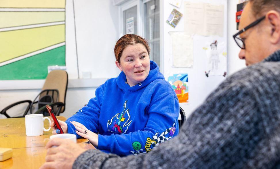 A woman is a blue hoodie is sitting at a table taking to a man. She is holding a mobile phone and there is a mug in front of her.