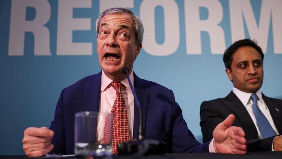 Nigel Farage in a blue suit and red tie makes a point at a Reform UK press conference. Reform UK chairman Zia Yusuf in a dark suit and blue tie sits next to him, looking to the right