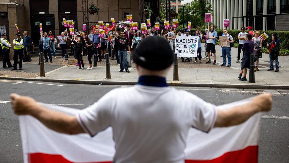 A man holds a flag bearing the St George's cross opposite protesters attending a rally organised by Stand Up To Racism outside the Britannia International Hotel on July 25, 2025 in London, England. 