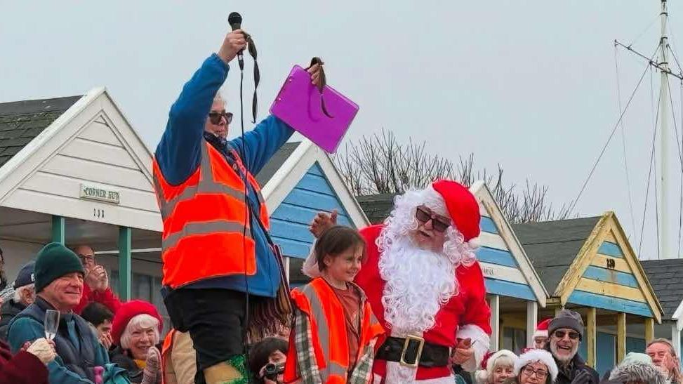 Reuben stands on a podium with a man dressed as santa clause. A volunteer in an orange hi-vis jacket stands on Reuben's other side and holds up his cut locks to the crowd