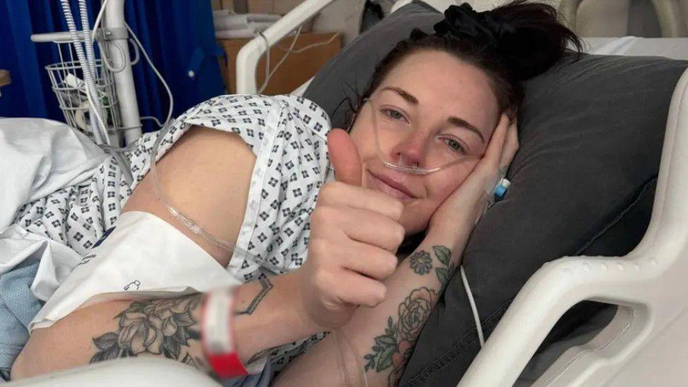 Lauren Lane in a hospital bed, surrounded by tubes and posing with a thumbs-up gesture