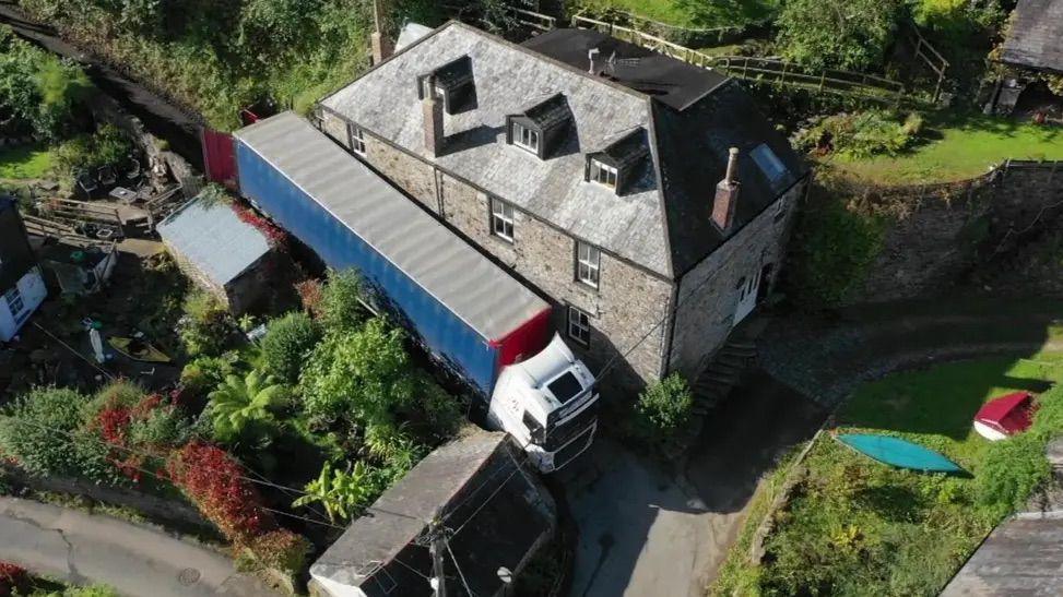 A lorry, in between a house and a wall. The back of the lorry doors are open. There are gardens around the road. 