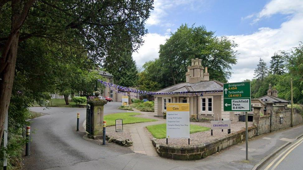 The entrance to a hospice site. There is a yellow and white branded signage, and a footpath leading to a single-storey building. The entrance is set back from the road, which has double-yellow lines running along it.