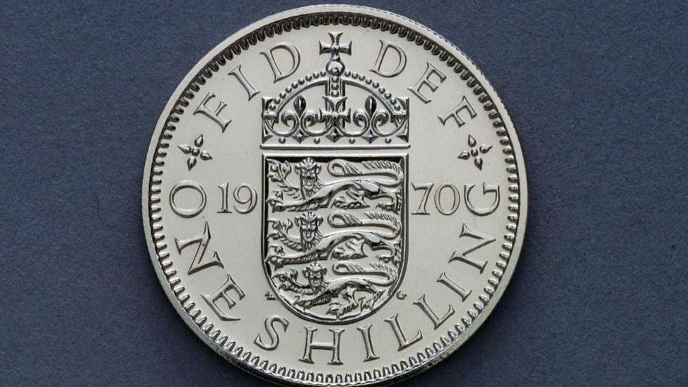 The shilling - a silver coin on a blue background. It says it is one shilling and had the three lions coat of arms on it. 