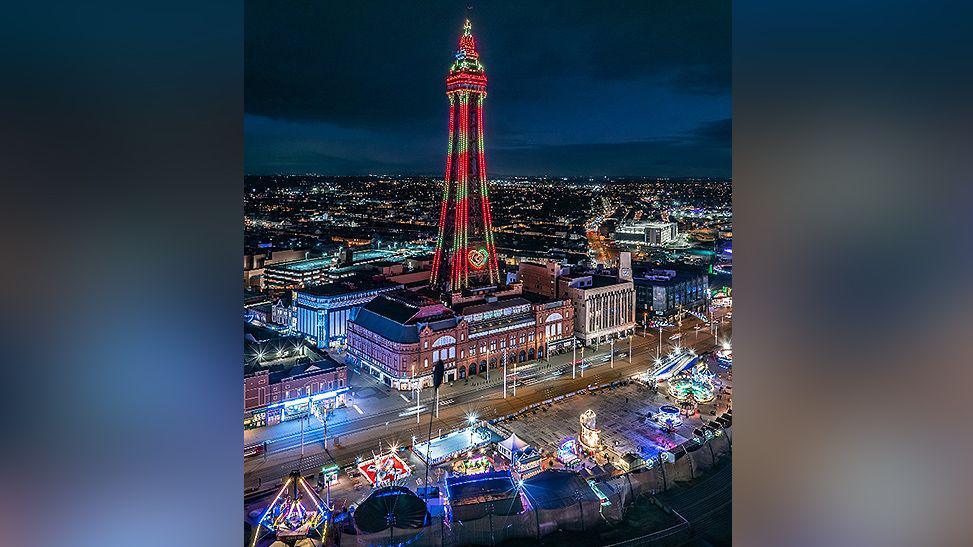 Aerial image of an illuminated Blackpool Tower and the illuminations at night. The Christmas by the Sea festival village is lit up on the other side of the Promenade.
