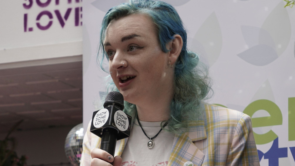 Ash Jones holding a microphone that says "show some love", wearing a pink, lilac and yellow tartan jacket