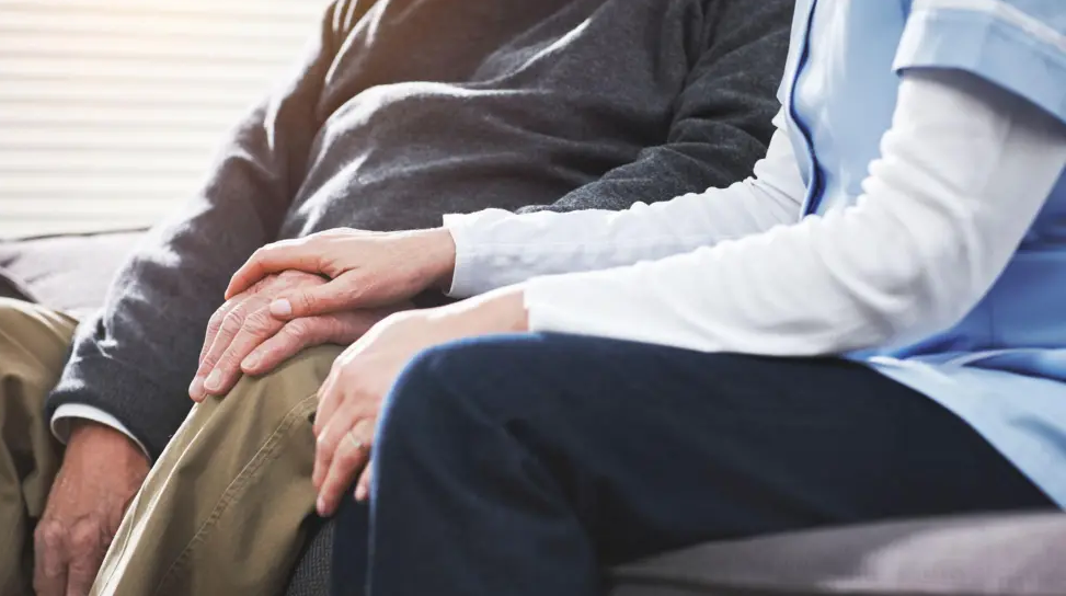 A stock picture of a nurse placing her hand on the hand of an elderly man. They are sitting on a sofa. Both are anonymous. 