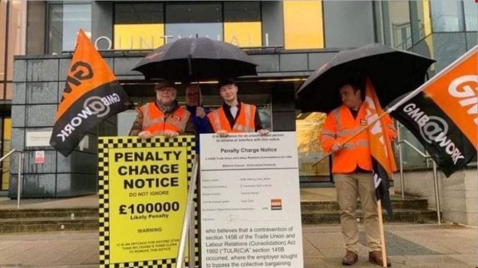 Three men in orange hi vis vests holding a placard 