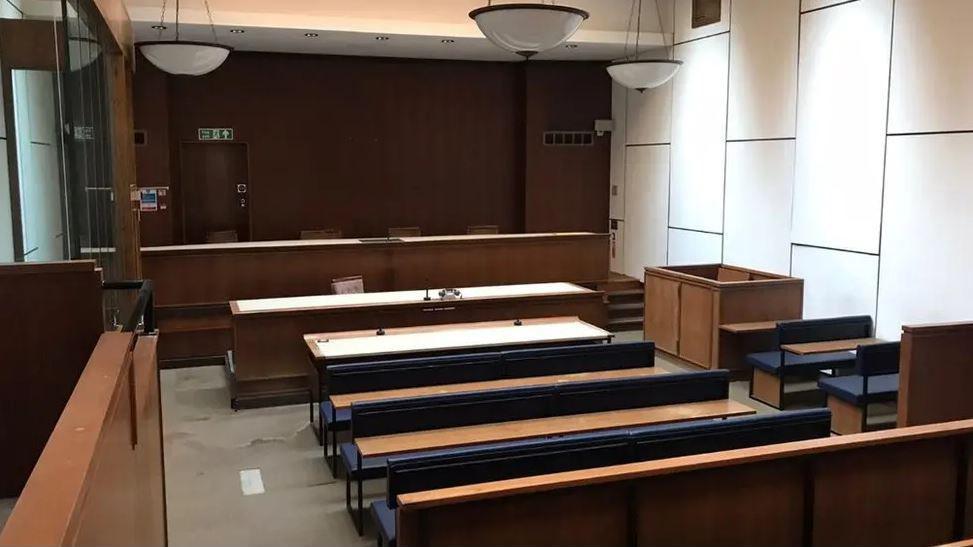 The inside of one of the empty courtrooms at Cirencester. The courtroom has a high ceiling, white walls and wooden desks