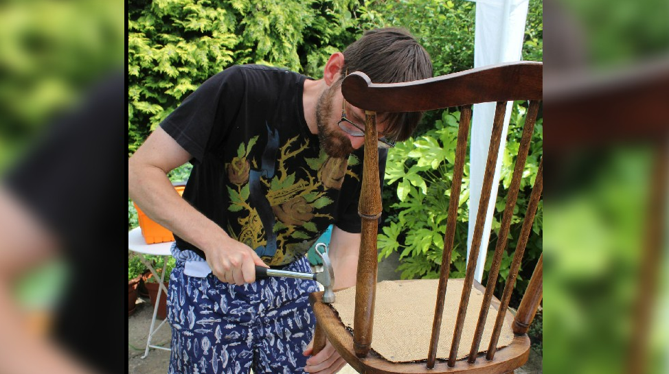 A man with a beard nailing studs into a dining chair, to secure a cushion.