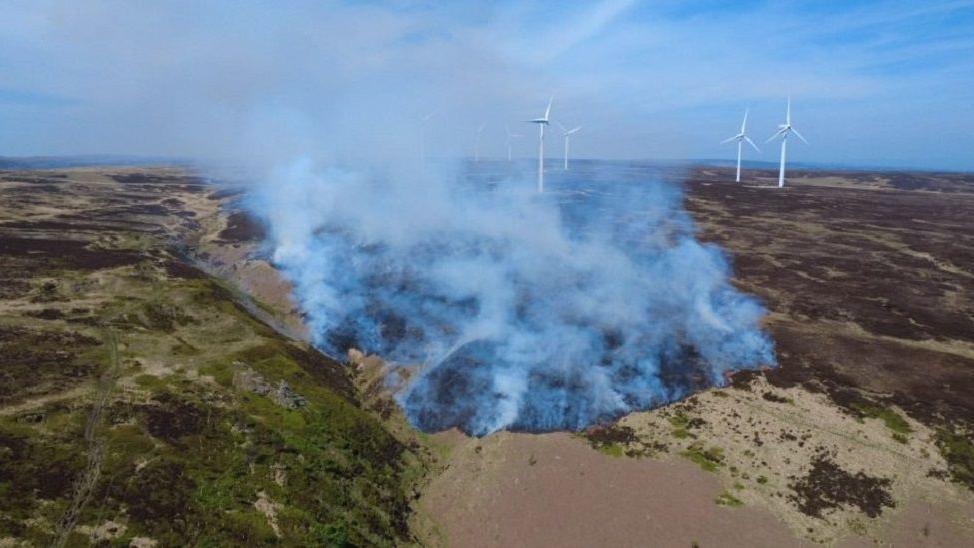 A drone shot of the wildfire, with white smoke billowing from green and brown grass and countryside. Solar powered windmills can also be seen in the background.