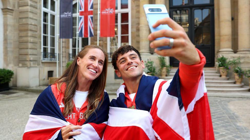  Helen Glover and Tom Daley of Team Great Britain pose for a selfie wearing their national flags during the Team Great Britain Flagbearer photocall at British Embassy on July 24, 2024 in Paris.