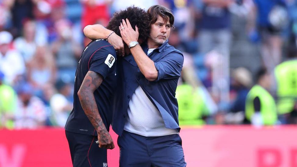 Pochettino has his arm around the neck of Richards who  has buried his head in the coach's shoulder. Pochettino's left hand is on the back of Richards' head as it to console him. The two men are on the football pitch and the crowd in the background are out of focus