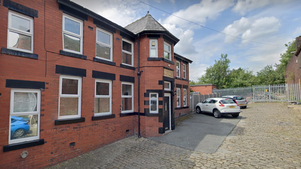 A street view image of an empty old building with orange brickwork. There are two cars outside in the car park.