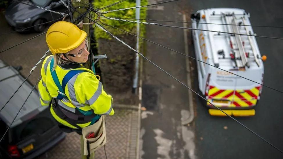 A female telecoms engineer is photographed fixing telegraph wires at the top of a pole. You can see the KCOM branded van parked on the road below.