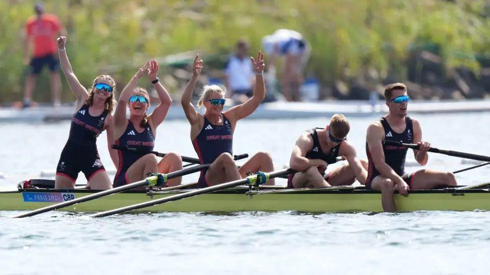 Five rowers celebrate in their boat after winning their final at the Paris 2024 Paralympics. The crew of three women have their arms aloft, while the two men at the rear of the boat are facing downwards as if recovering from their efforts.