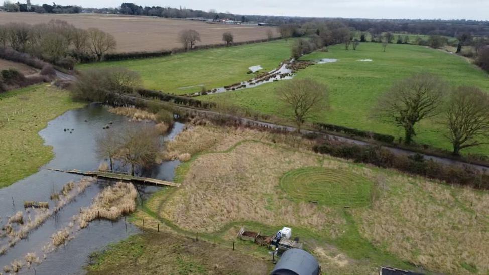 A picture of landscape from up high. There are fields in the back ground. In the foreground the you can see an area is flooded which is following the old river channel. There is a small bridge and you can see river wants to meander off into the distance. 