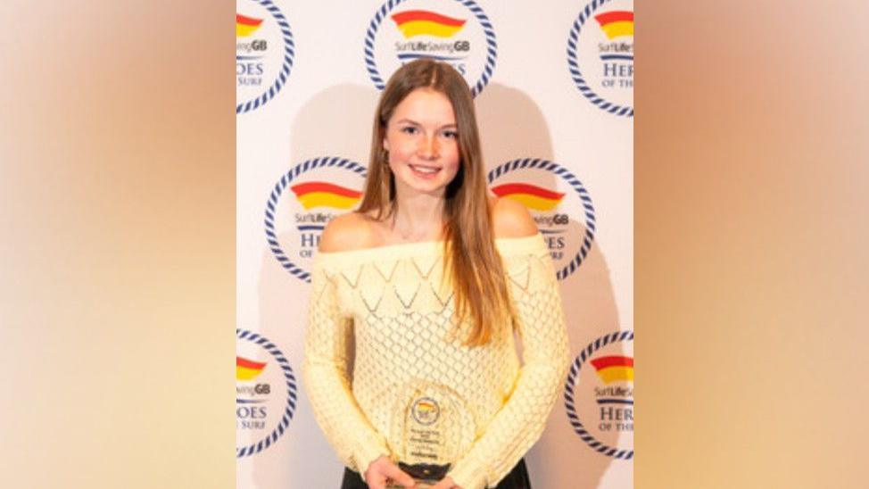 Esme Deacon holding her glass award in both hands at hip level. She is wearing a yellow top. She has long brown hair and is smiling at the camera. 