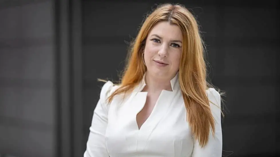 A headshot of Nicky Brennan, who has long red hair and is wearing a white blouse.