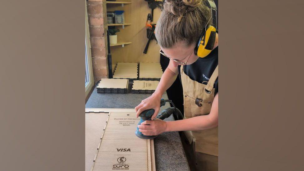 Yana at work using a sander on on of the box lids in her workshop. She is wearing yellow ear defenders and a stack of board pieces is in the background.
