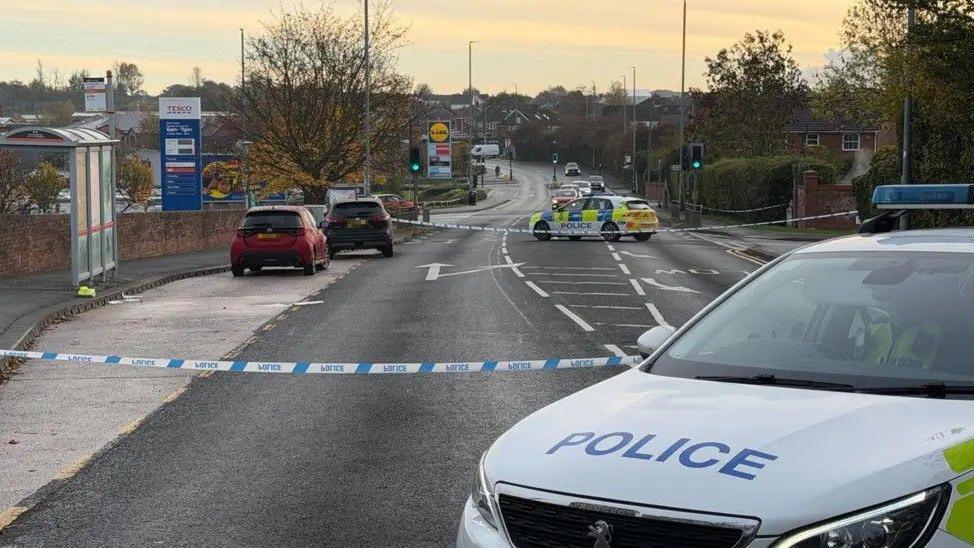 A police cordon in place along a suburban road. Visible are a bus stop, two parked cars within the cordon, two police cars and signs for Tesco and Lidl.