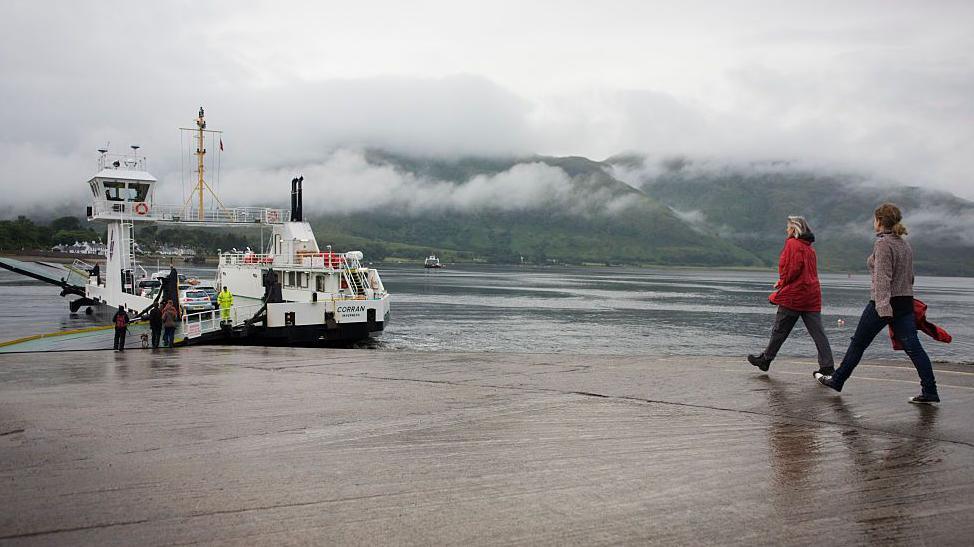 Two women walk towards the ferry, MV Corran. It is a wet day and the jetty is damp, and mist covers hills in the distance.