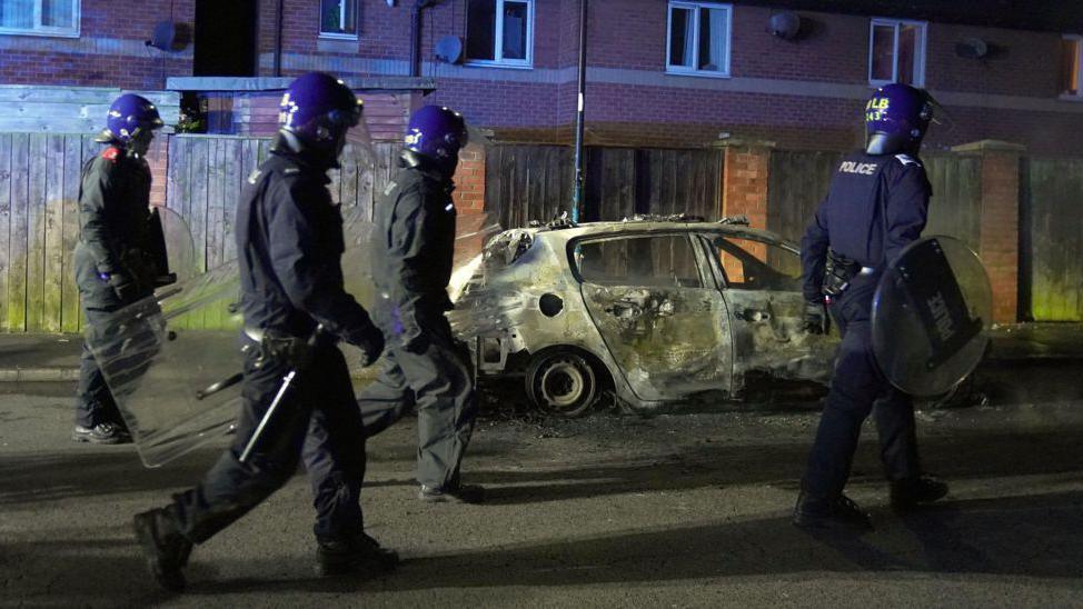 Four police officers wearing riot gear walking past a burnt-out car.