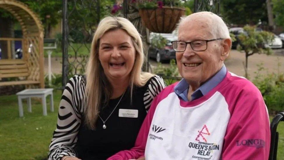 A man in a pink and white Queen's Baton Relay jumper smiles, sitting next to a blonce woman in a black and white shirt who is laughing. The pair are sitting in a garden with grass and benches in the background