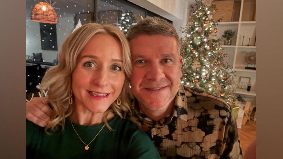 Claire has mid-length blonde hair and is wearing a green jumper and a gold pendant necklace. She is pictured with her husband Darren who is wearing a beige and black shirt and smiling at the camera. The couple are smiling at the camera while standing in front of a Christmas tree.