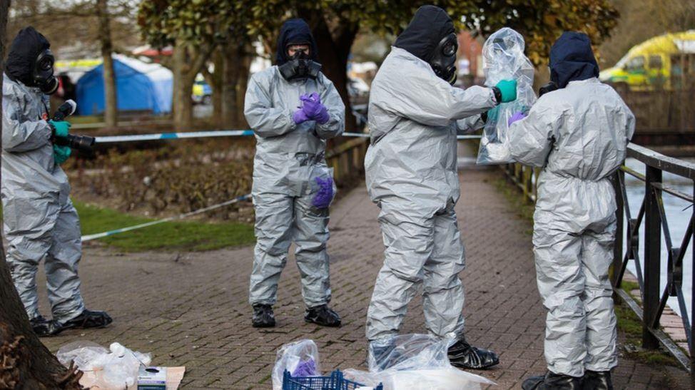 Officials in white protective suits and gas masks examine an area in Salisbury city centre next to a river. They are wearing purple gloves and putting evidence into clear bags. In the background a blue and white police tent is visible  as well as an ambulance.