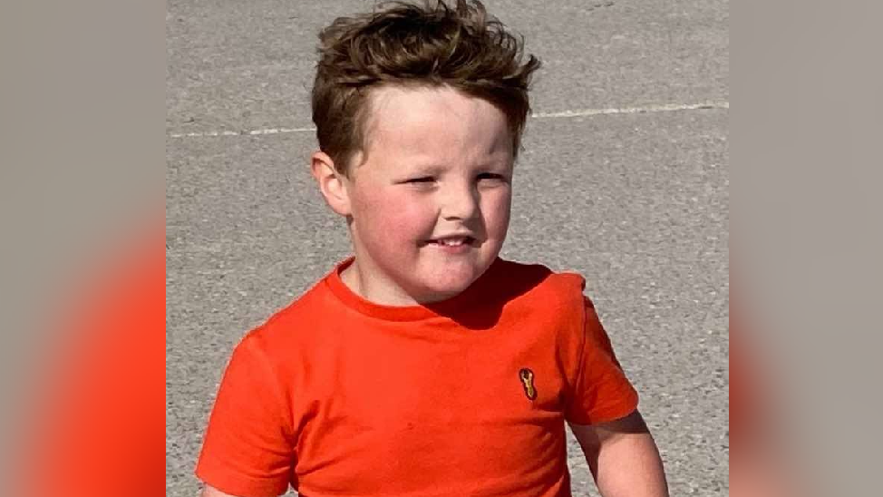 Ellis standing on grey concrete. He has short dark hair and is wearing a orange t-shirt. He is smiling and looking to the side.