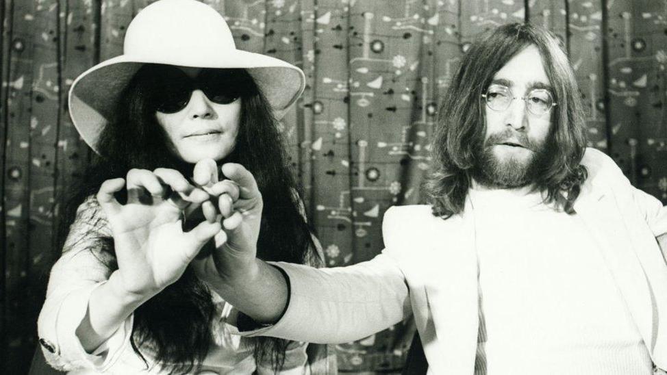 A black and white shot shows Yoko Ono, with long black hair, sunglasses and a white hat and partner John Lennon, with long dark hair, a beard and glasses, holding acorns towards the camera