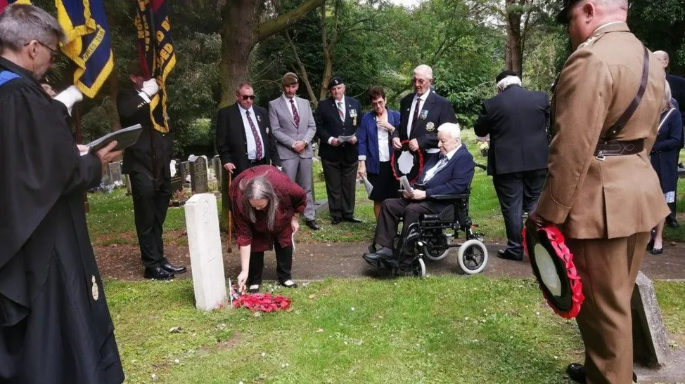 Several people gathered in a cemetery around a grave, including several men in military uniform. 