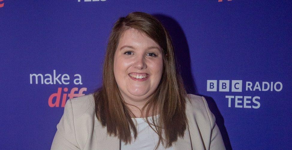 A head and shoulders shot of a smiling Rhiannon Matthews. She has long brown hair and is wearing a white top and cream blazer. The blue Make a Difference logo is in the background.