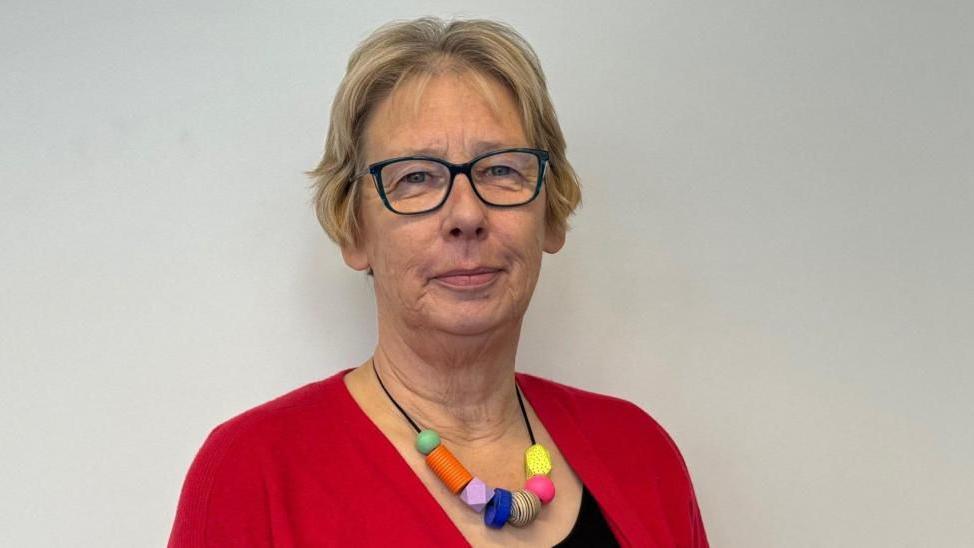 A woman with short blonde hair, glasses, a multi-coloured necklace, a red cardigan and black top, stands in front of a white wall looking at the camera.