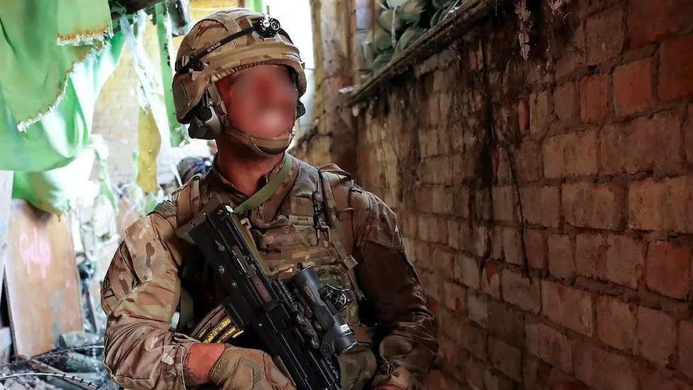 A Royal Marine wearing a helmet, body armour and carrying a gun looks up in a dilapidated building. The marine's face is blurred.