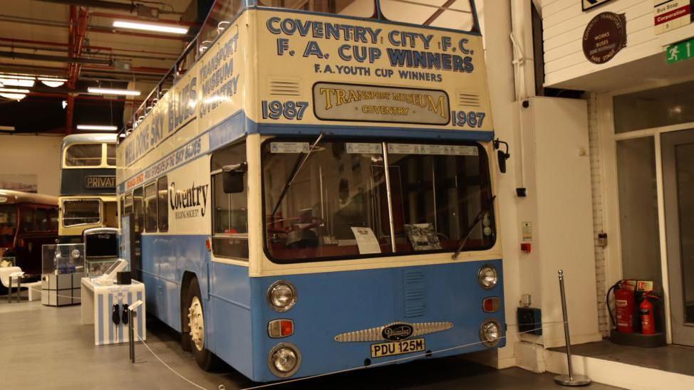 A blue and cream vintage bus, that open top and has writing on it that reads "Coventry City FC, FA Cup Winners. The bus is in a transport museum.