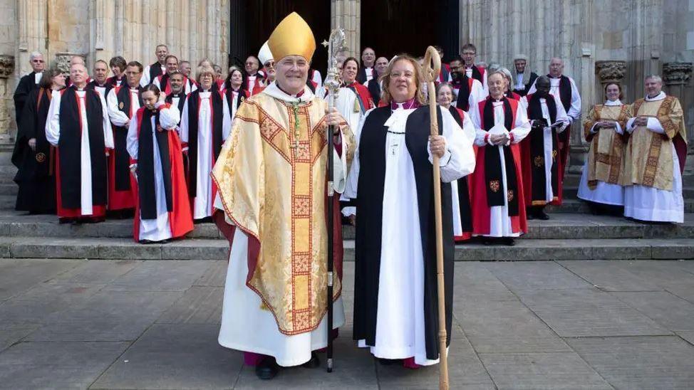 Venerable Patricia Hillas standing next to the Archbishop of York Stephen Cottrell outside York Minster. They are both in clerical dress and she is holding a staff. Other members of the clergy are lined up on the steps behind them.