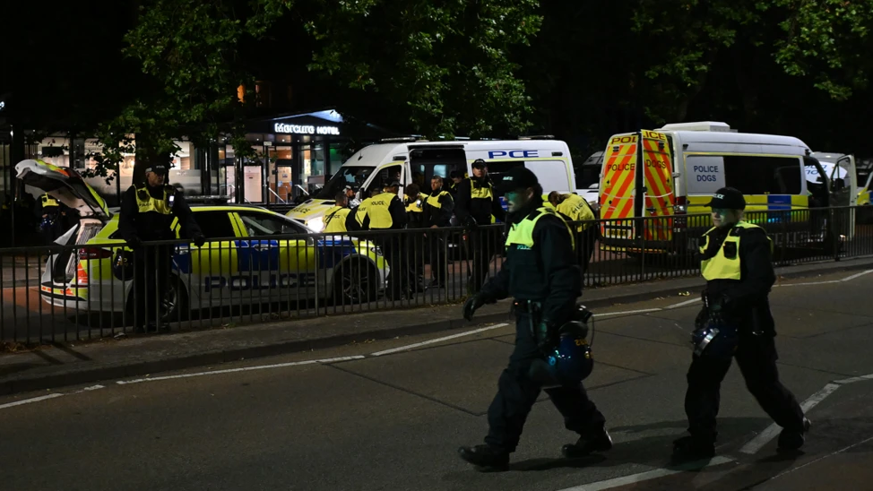 Police walking down the road in Bristol in riot gear. In the back of the image two police vans and a police car are parked in the road.
