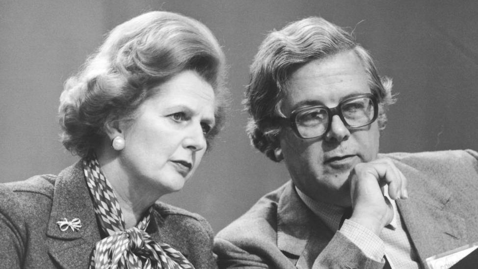 Black and white photograph of Margaret Thatcher sitting next to Geoffrey Howe at the Conservative Party Conference in Brighton, in October 1980.
She is wearing a neckerchief and a brooch. Howe is wearing glasses and is resting his chin on his hand.