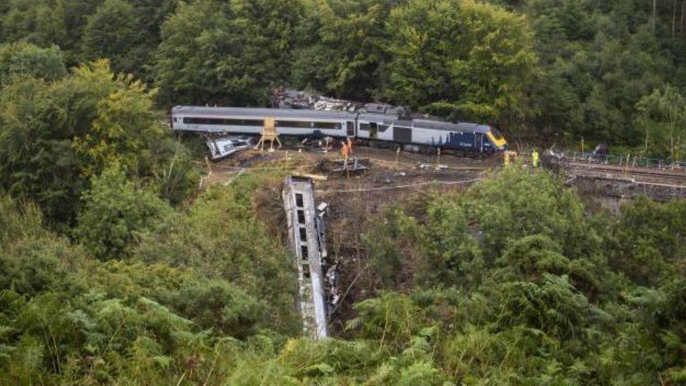 Stonehaven derailment - image of a train on tracks in woodland, and also a burned carriage down a slope.