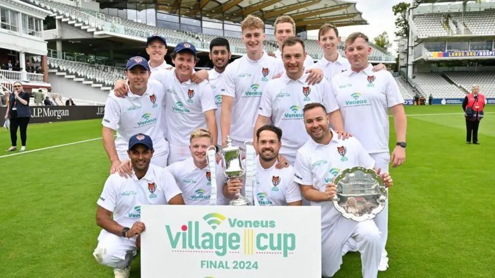 A team line-up featuring 13 men wearing cricket whites. One man holds a silver cup, another a silver platter and a third a placard saying "Voneus Village Cup Final 2024".