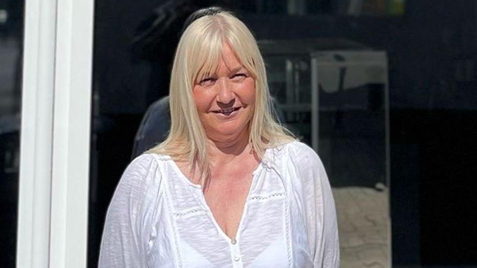 A woman with mid length blonde hair and wearing a white long sleeved top is stood i the doorway of a shop.