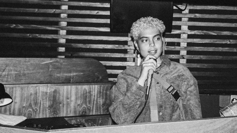 Marley Small is standing in a DJ booth speaking into a microphone. She is wearing a jacket and has short hair. The picture is black and white.