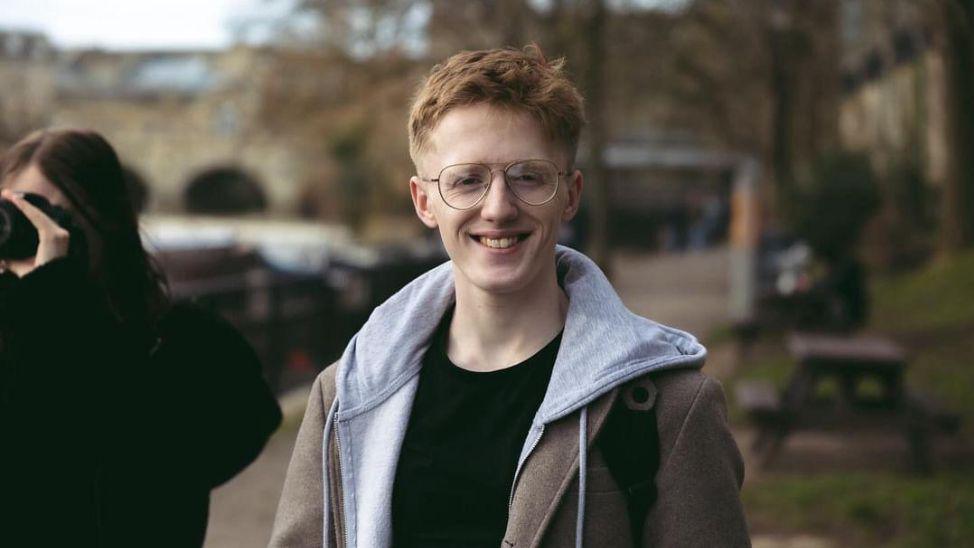 A man wearing glasses, a grey hooded top and a brown coat, looking directly at the camera. He is stood in a park. 