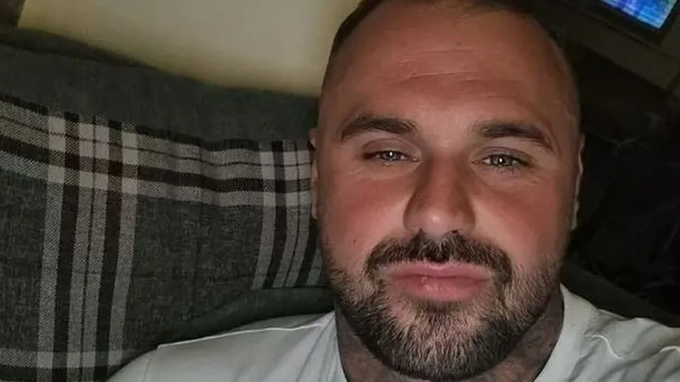 A selfie of Tom Gaughan. He is sitting on a grey, black and white sofa and smiling into the camera. He has blue eyes and a dark beard.