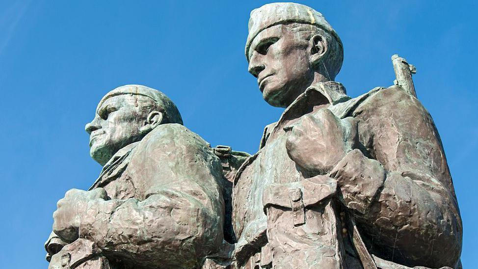 A close-up of two figures in the Commando Memorial near Spean Bridge. The men are portrayed wearing military fatigues and headwear. They are equipped with webbings, back packs and rifles. The figures are pictured against a blue sky.